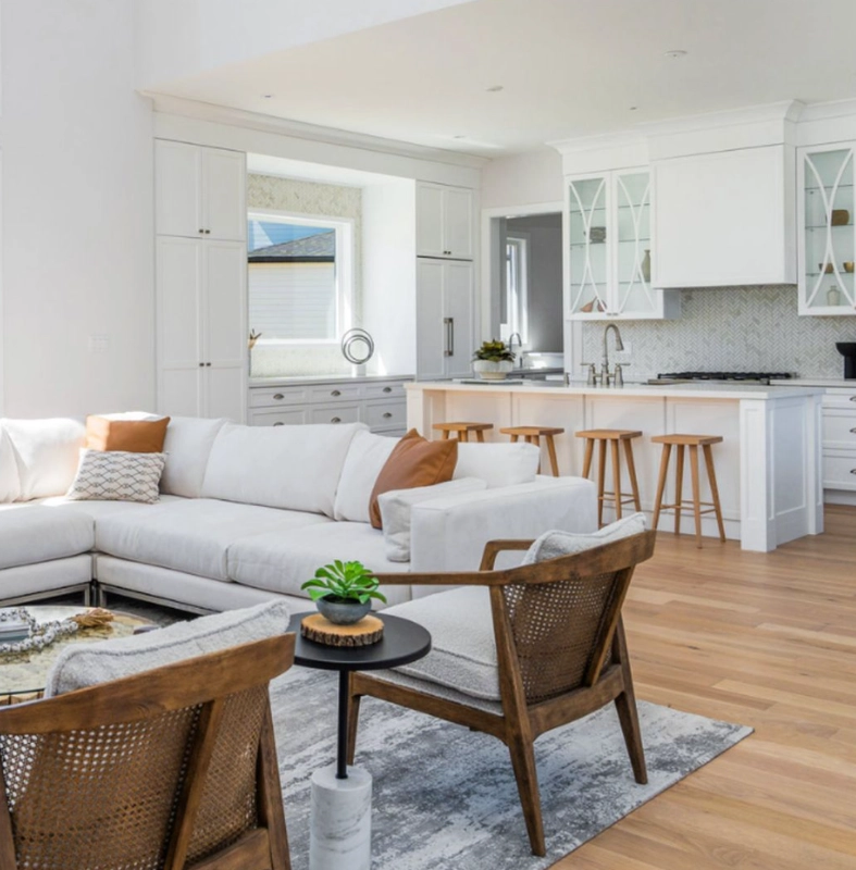 An open-plan living room and white kitchen with warm wood floors and woven accent chairs.